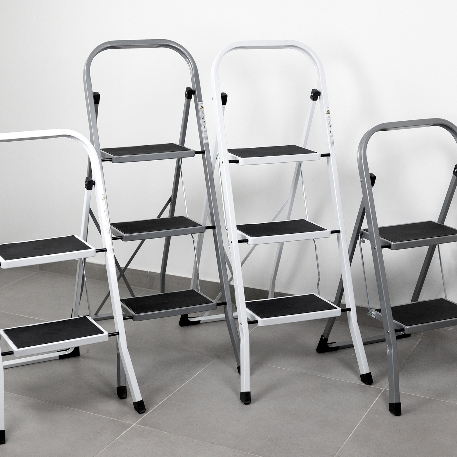 Mehrere Klappleitern stehen auf einem grauen Boden vor einer weißen Wand