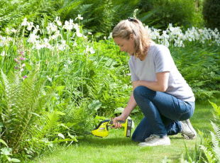 Eine Frau kniet im Garten und schneidet Pflanzen mit einem kleinen elektrischen Trimmer