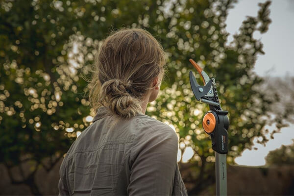 Eine Person mit zusammengebundenem Haar steht im Garten und hält eine Astschere
